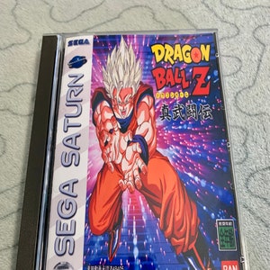 Puede incluir: Un cartucho de videojuego Sega Saturn para Dragon Ball Z: Real Bout. El juego presenta una imagen colorida de Goku en su forma Super Saiyan sobre un fondo morado y azul. El texto en el cartucho dice "Dragon Ball Z" y "Real Bout".