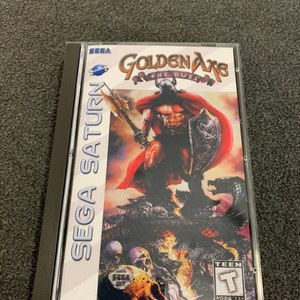 Puede incluir: Caja de juego de Sega Saturn para Golden Axe: The Duel. La portada presenta a un guerrero musculoso con armadura roja sosteniendo un hacha grande y un escudo, de pie en un paisaje de fuego con calaveras y huesos. El juego está clasificado como Teen por la ESRB.