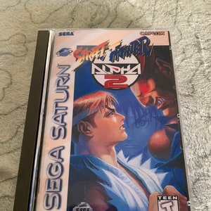 Może przedstawiać: Pudełko gry Sega Saturn do Street Fighter Alpha 2, z niebiesko-czerwoną ilustracją dwóch walczących postaci. Pudełko jest czarne z przezroczystym plastikowym okładką i tekstem "SEGA SATURN" nadrukowanym pionowo po lewej stronie. Klasyfikacja ESRB to "TEEN", a gra jest licencjonowana przez Sega Enterprises Ltd.