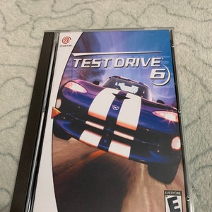 Op de afbeelding: Een Dreamcast-gamecase voor Test Drive 6. De cover toont een blauwe sportwagen met witte racestrepen. De speltitel is wit en het Sega-logo is zichtbaar. De case zit in een doorzichtige plastic hoes.