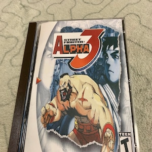 May include: A Sega Dreamcast game case for Street Fighter Alpha 3. The case is white with a red and black design. The game features a muscular character with a red and black outfit. The ESRB rating is Teen.