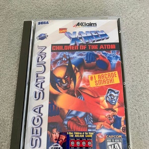 Puede incluir: Una caja de juego de Sega Saturn para X-Men: Children of the Atom. La caja es azul y negra con una imagen de Wolverine y Cíclope luchando. El juego está clasificado como K-A para niños y adultos por Capcom.