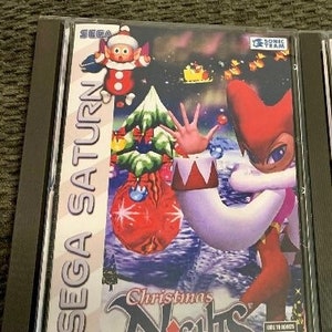 Puede incluir: Caja de juego de Sega Saturn para Christmas Nights into Dreams. La caja es blanca con detalles rojos y azules. La parte delantera de la caja presenta un personaje de dibujos animados con ropa roja y blanca. El juego está clasificado como "KA" para niños y adultos.