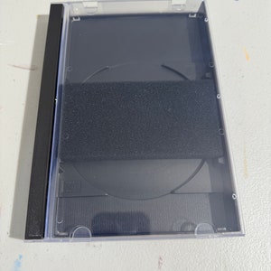 Puede incluir: Una caja de CD de plástico transparente con una columna vertebral negra y un inserto de espuma negra para sujetar el CD. La caja está diseñada para contener un CD estándar.