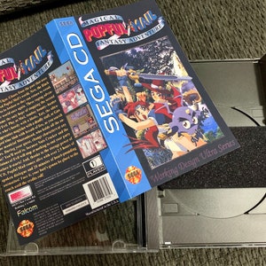 May include: A Sega CD game case for "Magical Pop'n Mail: Fantasy Adventure" is open, revealing the game's CD and packaging. The box art features colorful anime-style characters. The case is silver with a black foam insert.