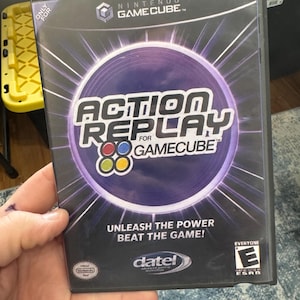 Pode incluir: Uma caixa de jogo preta da Nintendo GameCube Action Replay com um gráfico circular roxo e texto branco que diz Action Replay for GameCube, Unleash the Power Beat the Game, um logótipo Datel e um símbolo de classificação ESRB Everyone.
