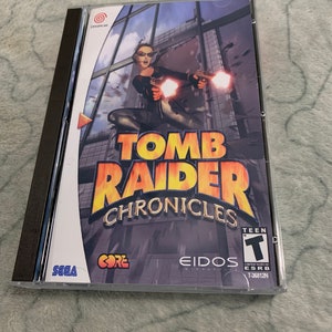 Puede incluir: Una caja de juego Sega Dreamcast para Tomb Raider Chronicles. La caja es de plástico transparente con una columna vertebral negra. La parte frontal de la caja presenta a una mujer con un atuendo negro sosteniendo dos pistolas. El título del juego está en letras grandes amarillas con un contorno rojo. La clasificación PEGI es 12.