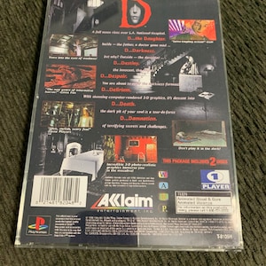 Puede incluir: Caja de juego de PlayStation para "D", un juego de terror. La parte trasera de la caja presenta un diseño oscuro con imágenes y texto, incluyendo "D...the Daughter" y "Don't play it in the dark!" El juego está clasificado como Teen por sangre y violencia animadas.