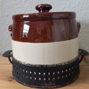 May include: A vintage ceramic crock with a brown top and a cream-colored midsection. It has a matching lid and sits on a black metal stand. The crock is likely used for food storage.