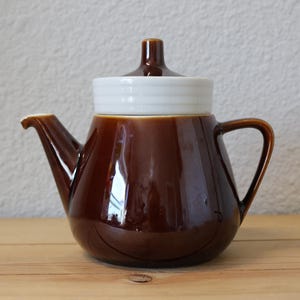 May include: A brown ceramic teapot with a white lid. The teapot has a rounded body and a spout on one side and a handle on the other.