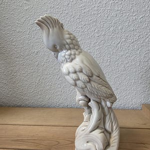 May include: A white ceramic cockatoo figurine perched on a decorative base. The bird has detailed feather textures and a prominent crest. The figurine is a neutral color and sits on a wooden surface.