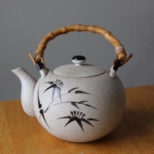 May include: A white ceramic teapot with a bamboo handle and black floral design. The teapot has a lid and a spout.