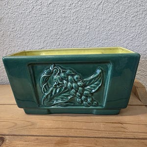Puede incluir: Una jardinera rectangular de cerámica verde oscuro con un diseño en relieve de uvas y hojas en la parte delantera. El interior es de color amarillo brillante. La jardinera está sobre una superficie de madera contra una pared texturizada.