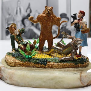 May include: A detailed figurine depicting characters from The Wizard of Oz. The Lion, Tin Man, Scarecrow, Dorothy, and Toto are arranged on a yellow brick road base, set on a marble-like pedestal. The figures are painted in vibrant colors.