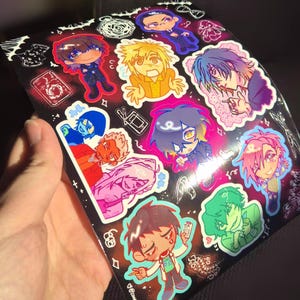 May include: A sheet of colorful anime-style stickers featuring various characters with different hair colors and outfits. The stickers are on a black background with white and red accents. The stickers are held by a hand.