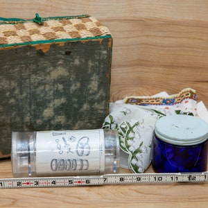 May include: A wicker basket with a green lid, a clear plastic container with a label showing a diagram of a set of nesting bowls, a blue glass jar with a white lid, and a white fabric with a floral pattern.