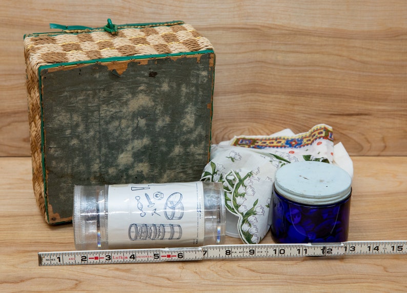 May include: A wicker basket with a green lid, a clear plastic container with a label showing a diagram of a set of nesting bowls, a blue glass jar with a white lid, and a white fabric with a floral pattern.
