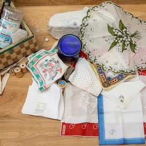 May include: A wicker basket filled with sewing supplies, including spools of thread, buttons, and needles. There are also several handkerchiefs with floral patterns and embroidery, and a blue glass jar with the word "Foxzema" on it.