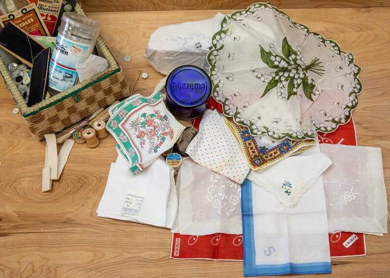 May include: A wicker basket filled with sewing supplies, including spools of thread, buttons, and needles. There are also several handkerchiefs with floral patterns and embroidery, and a blue glass jar with the word "Foxzema" on it.