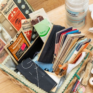 May include: A vintage sewing box filled with various sewing supplies, including needles, buttons, thread, and fabric scraps. The box is made of wicker and has a floral design. The box is open and the contents are visible. The text on the packaging reads "Self Cover Buttons Quarter Ball Style" and "7 Home Repair Needles".