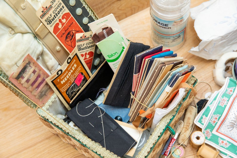 May include: A vintage sewing box filled with various sewing supplies, including needles, buttons, thread, and fabric scraps. The box is made of wicker and has a floral design. The box is open and the contents are visible. The text on the packaging reads "Self Cover Buttons Quarter Ball Style" and "7 Home Repair Needles".