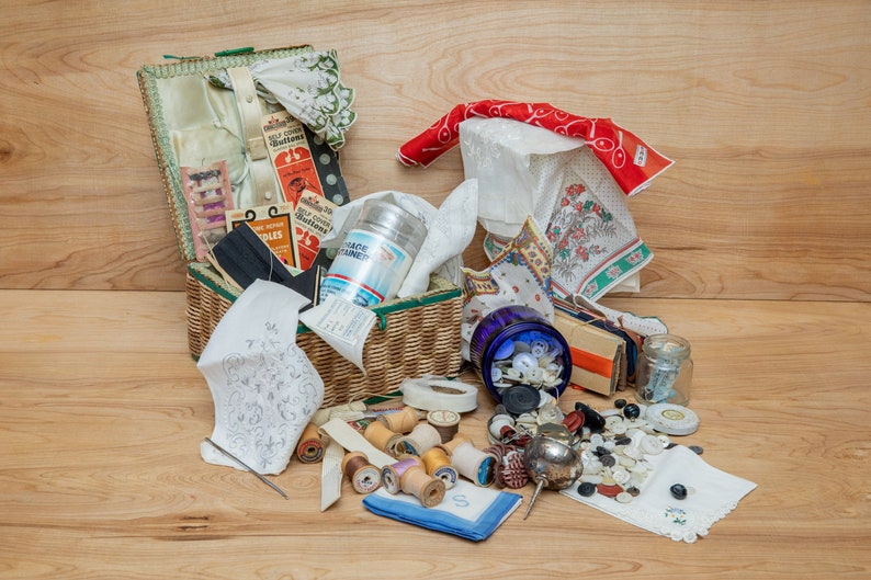 May include: A wicker basket filled with sewing supplies, including spools of thread, buttons, needles, and fabric scraps. The basket is surrounded by additional sewing supplies, including a jar of buttons, a spool of ribbon, and a small jar of needles.