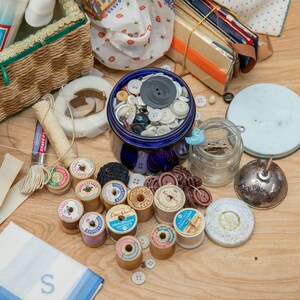May include: A collection of sewing supplies including spools of thread, buttons, and a jar of pins. The supplies are arranged on a wooden surface.