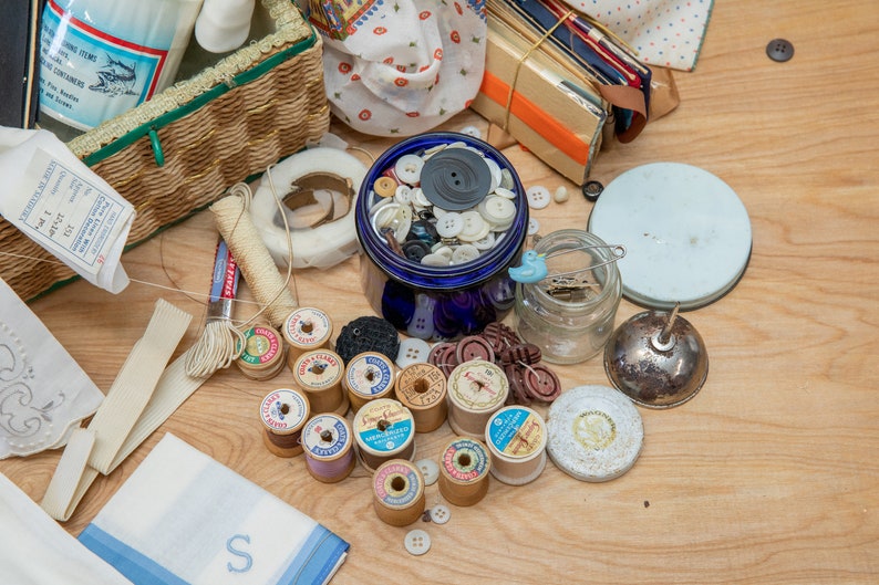 May include: A collection of sewing supplies including spools of thread, buttons, and a jar of pins. The supplies are arranged on a wooden surface.