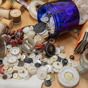 May include: A blue glass jar filled with white and blue buttons spills onto a wooden surface. The buttons are scattered around a variety of other sewing supplies, including spools of thread, a metal thimble, and a small glass jar.