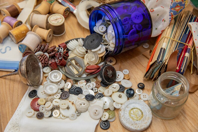 May include: A blue glass jar filled with white and blue buttons spills onto a wooden surface. The buttons are scattered around a variety of other sewing supplies, including spools of thread, a metal thimble, and a small glass jar.