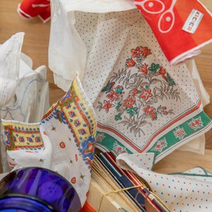May include: A collection of vintage handkerchiefs with floral and geometric patterns in shades of red, blue, green, and white. The handkerchiefs are arranged on a wooden surface with a blue glass jar and a wicker basket.