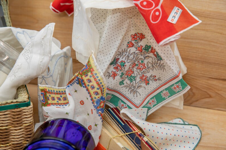 May include: A collection of vintage handkerchiefs with floral and geometric patterns in shades of red, blue, green, and white. The handkerchiefs are arranged on a wooden surface with a blue glass jar and a wicker basket.