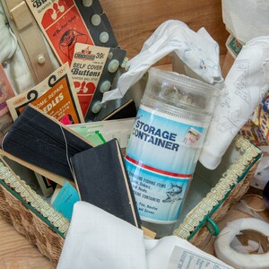 May include: A wicker basket filled with various sewing supplies, including needles, thread, buttons, and fabric scraps. A clear plastic container labeled "STORAGE CONTAINER" is visible in the center of the image. The basket is sitting on a wooden surface.