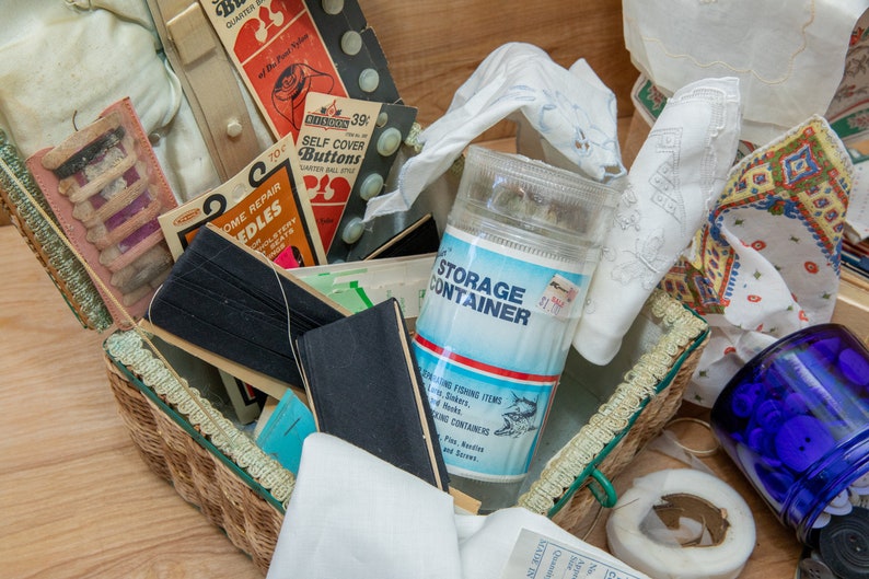 May include: A wicker basket filled with various sewing supplies, including needles, thread, buttons, and fabric scraps. A clear plastic container labeled "STORAGE CONTAINER" is visible in the center of the image. The basket is sitting on a wooden surface.