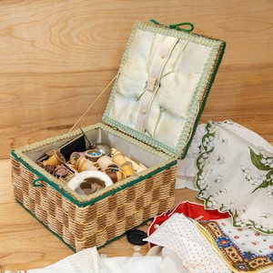 May include: A wicker sewing basket with a green and white lid. The basket is filled with spools of thread, buttons, and other sewing supplies. The lid is lined with a white fabric and has a green trim.