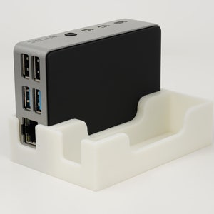 May include: A small, silver computer with a black top, sitting in a white plastic stand. The computer has several ports on the side, including USB and ethernet ports.