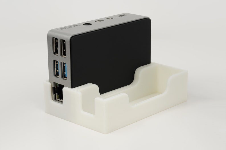 May include: A small, silver computer with a black top, sitting in a white plastic stand. The computer has several ports on the side, including USB and ethernet ports.