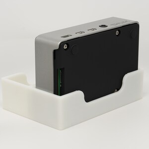 May include: A silver and black device with a white plastic stand. The device has a small green circuit board visible on the side. The device has a small black button and a small black circle on the back. The device has a small black text that says 'Smart' on the back.
