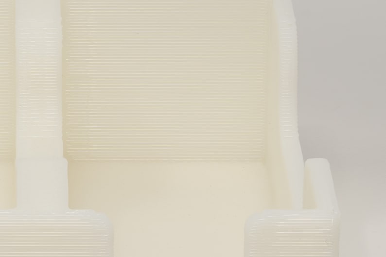 May include: Close-up of a white plastic object with a textured surface. The object has a rectangular shape with two open sides. The object is likely a 3D printed model.