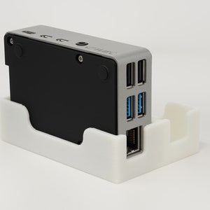 May include: A small, silver and black computer device with multiple ports, including USB, Ethernet, and audio. The device is sitting in a white plastic stand.