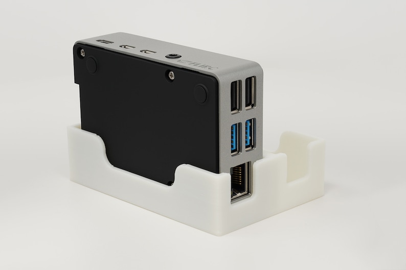 May include: A small, silver and black computer device with multiple ports, including USB, Ethernet, and audio. The device is sitting in a white plastic stand.