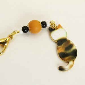 May include: A gold-colored lobster clasp connected to a beaded strand and a cat charm. The charm is a calico cat with black, brown, and white markings. The beads are black, white, and a large tan bead.