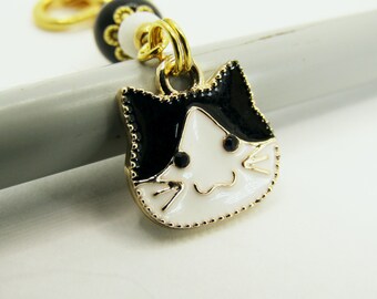 May include: A gold-toned cat charm with black and white enamel details. The cat's face is white with black accents and a smiling expression. The charm is attached to a gold chain and a black and white bead.