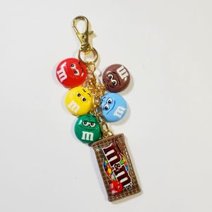 May include: A gold-toned metal keychain featuring five colorful M&M character charms in red, yellow, green, blue, and brown, each with a white letter m. A rectangular charm shaped like a chocolate bar with the M&M logo is attached to the chain.