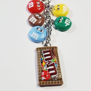 May include: A metal keychain featuring five colorful M&M character charms in red, brown, light blue, yellow, and green, attached to a silver chain. A rectangular charm shaped like a chocolate bar package with the M&M logo is also attached.