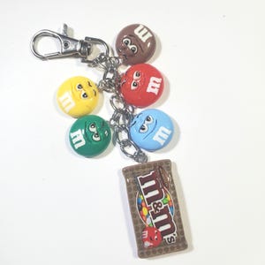 May include: A silver-toned metal keychain featuring a lobster clasp and a chain with five attached charms. The charms include four round M&M character faces in brown, red, yellow, green, and blue, plus a rectangular M&M milk chocolate bar charm.