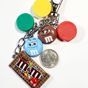 May include: A metal keychain featuring a rectangular M&M's milk chocolate candy bar charm, a blue M&M character charm, a brown M&M character charm, and three solid circular charms in yellow, green, and red, displayed next to a US dime coin.