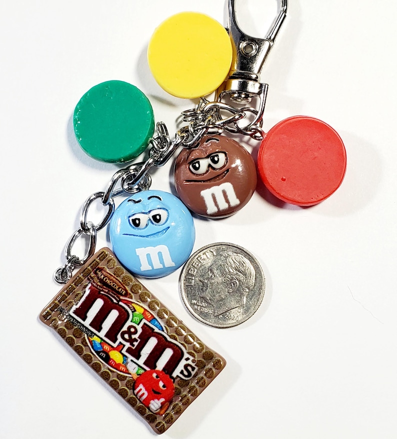 May include: A metal keychain featuring a rectangular M&M's milk chocolate candy bar charm, a blue M&M character charm, a brown M&M character charm, and three solid circular charms in yellow, green, and red, displayed next to a US dime coin.