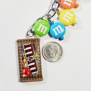 May include: A silver-toned metal keychain featuring a brown M&M's milk chocolate bar charm and several round, colorful M&M's-themed beads in green, blue, yellow, and orange. A U.S. dime is placed next to the charm to provide a scale reference.