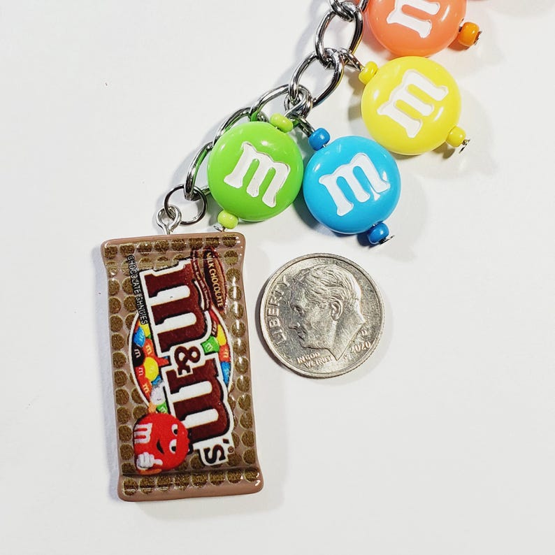 May include: A silver-toned metal keychain featuring a brown M&M's milk chocolate bar charm and several round, colorful M&M's-themed beads in green, blue, yellow, and orange. A U.S. dime is placed next to the charm to provide a scale reference.
